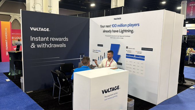In this image, two people can be seen at a trade show booth for a technology company called "Voltage". They appear to be having a conversation with another person who has a laptop open. The booth features an interactive display showcasing the lightning network that powers many online services such as email and social media platforms like Facebook. There is also a sign on the booth that promotes instant rewards and free currency trading.