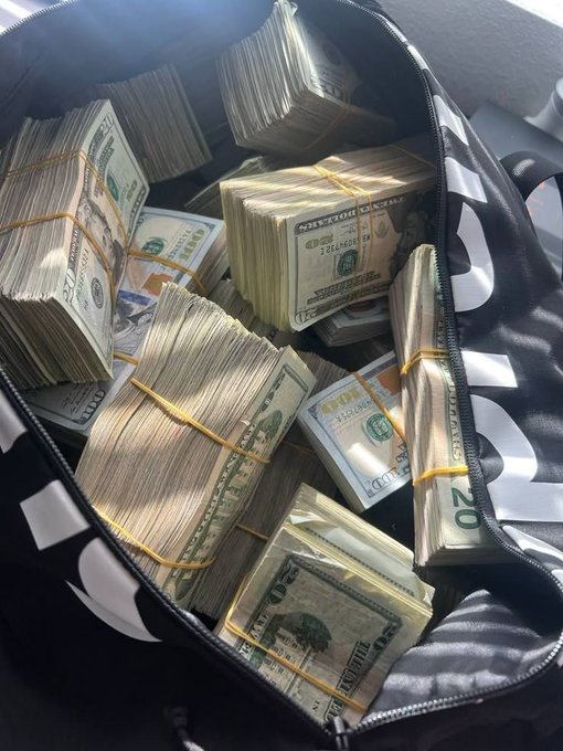 An open black and white bag filled with numerous thick stacks of US twenty-dollar bills, secured with rubber bands.
