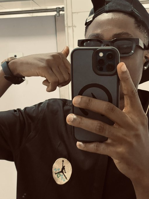 A person takes a mirror selfie with their iPhone, wearing a black t-shirt featuring a Jordan logo, a black cap, and dark sunglasses, while giving a thumbs-up gesture.