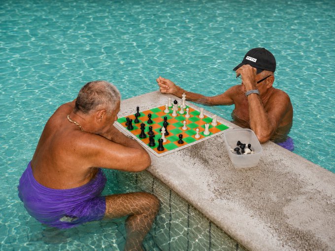Two shirtless men in a swimming pool play chess on a makeshift board set atop a ledge, with one wearing a black cap reading "RETIRED"—a fitting visual metaphor for the post's claim that even the "retired" Pyth team is actively driving massive RWA volume growth, suggesting the crypto market data sector is still in its early innings despite already hitting $110B in monthly volume.