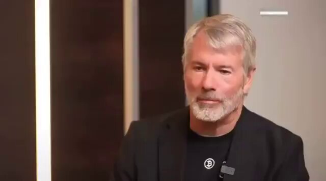 The image features a man wearing a black suit jacket with a white collar sitting against a brown wall. The man has grey hair and a beard, giving him an older appearance. He appears to be looking directly at the camera while seated on a chair in front of a desk. There is also text visible in the image that reads "Focus on what is in the image and how it relates to the post: 'Michael Saylor once said if Strategy sells bitcoin they will buy multiples more $BTC than they sold: even if we were to sell 1 bitcoin, we'd be buying 10-20 more bitcoin.'".