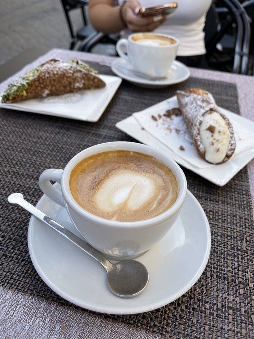 A cappuccino with heart-shaped latte art sits on a saucer next to a spoon, accompanied by two cannoli desserts on white plates at an outdoor café table.