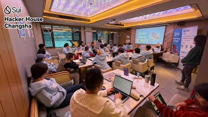 A group of around 20 developers work on laptops at the Sui Hacker House Changsha, with a presentation screen displaying "Sui" branding and the text "破浪长航 修远万里" visible in the background. The scene illustrates the post's narrative of 22 builders collaborating intensively in one house under mentors from Bucket, Cetus, and CCP Games.