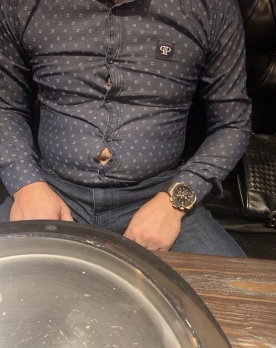 A photo of a man wearing a fitted navy blue patterned shirt with a 'TP' logo, showing his torso and a wristwatch, seated with an empty metal plate in the foreground.