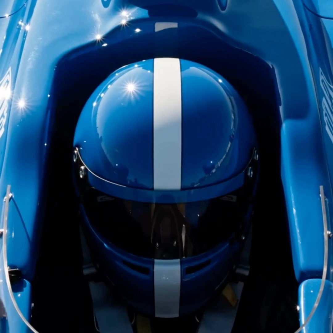 A close-up overhead view of a glossy blue race car helmet with a white center stripe, showing reflections of overhead lights on its surface.