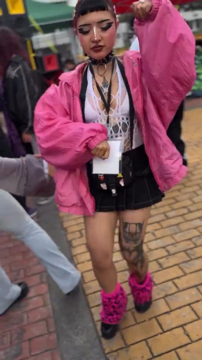A person with a septum piercing, gothic makeup, and a chain choker posing on a brick street, wearing an oversized pink jacket, white mesh top, black mini skirt, and fuzzy pink leopard-print boots, with decorative charms hanging from their skirt.