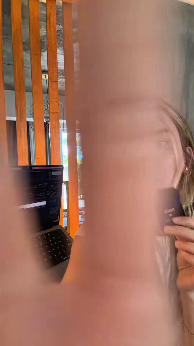 The image shows a woman holding a black device with a camera lens attached to it, possibly taking a picture of herself. The background features an orange wall with vertical stripes, which adds a unique visual element to the scene. A laptop can be seen in the background as well.