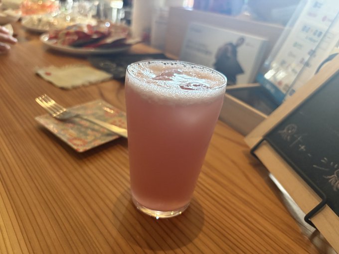 A glass of pink-colored beverage with foam sits on a wooden table next to a fork and decorative napkin, with blurred plates and dining items in the background.