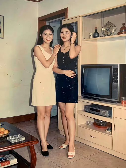 Vintage-style photograph of two young women in dresses posing next to a television set in a domestic living room setting.