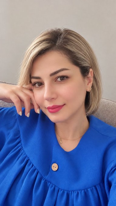 A close-up portrait of a smiling woman with short blonde hair, wearing a blue blouse and a small gold necklace, resting her chin on her hand.