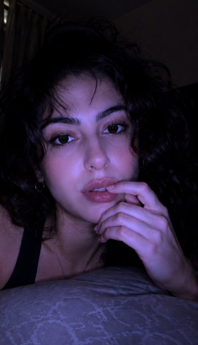 A close-up, dimly lit selfie of a young woman with curly dark hair, resting her fingers near her lips.