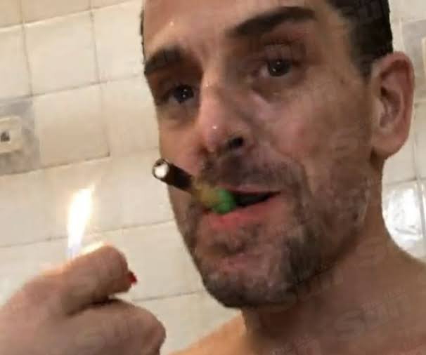 A close-up of a shirtless man with a goatee attempting to light a cigar that is already in his mouth.