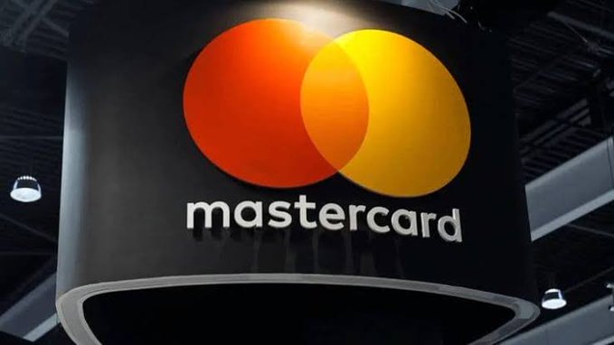A large, illuminated Mastercard sign featuring the company's iconic overlapping red and yellow circles logo is displayed at what appears to be a corporate or trade show venue. The image visually represents the post's point about Mastercard's expanding role in blockchain initiatives, including its growing engagement with the XRP Ledger.