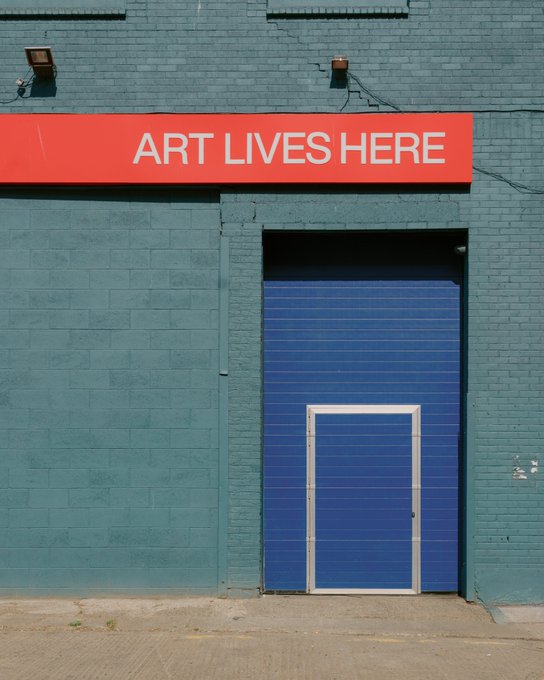 A blue door set within a teal-painted brick wall beneath a red sign reading "ART LIVES HERE," with a smaller white-framed door nested within the larger blue door. The image visually embodies the post's sentiment about art persisting in imperfect, weathered spaces—capturing the "crumbling facade" mentioned while the bold signage affirms that creativity and artistic life endure within.