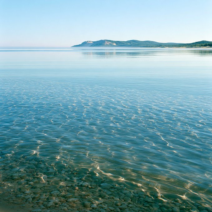 A serene, clear turquoise sea with gentle ripples, revealing smooth pebbles on the shallow seabed, stretches toward distant green hills under a pale blue sky.