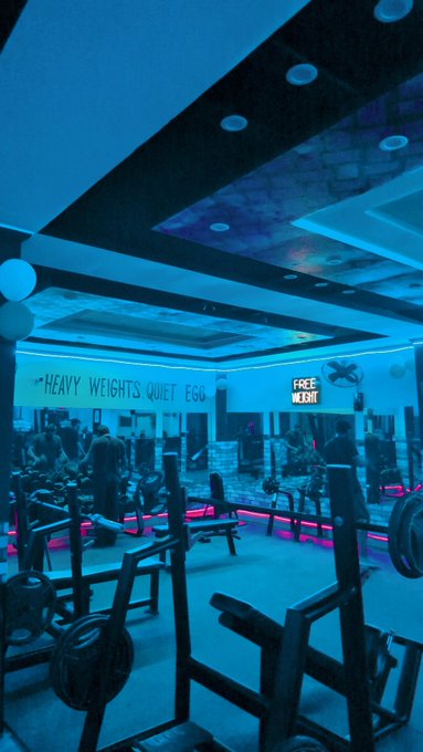 A gym interior with blue lighting, featuring weight racks, exercise equipment, and wall signage reading 'HEAVY WEIGHTS QUIET EGO' and 'FREE WEIGHT'.