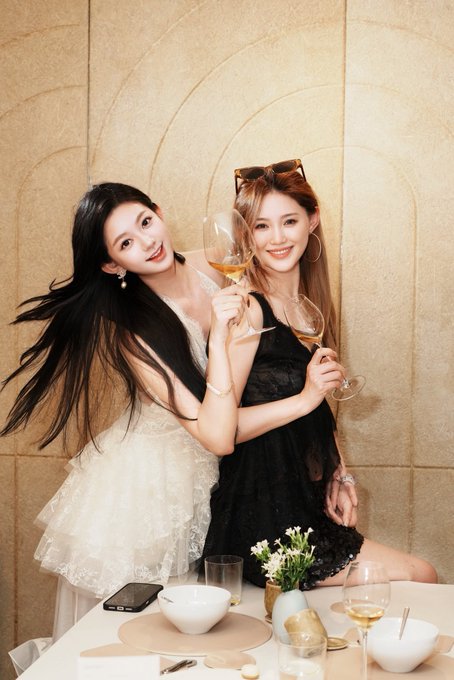Two smiling young women in elegant dresses (one white lace, one black) holding wine glasses at a table with dishes and flowers, posing for a stylish portrait.