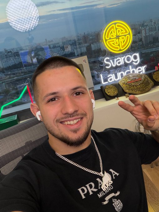 A smiling man wearing a black Prada t-shirt, chain necklace, and white earbuds poses indoors with championship belts visible behind him, with a neon sign reading 'Svarog Launcher' illuminated on the window.