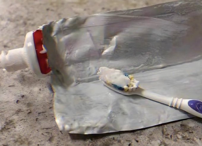 A close-up of a silver metallic pouch, likely a toothpaste tube, with a dollop of toothpaste squeezed onto a toothbrush resting on top of it, sitting on a speckled stone surface.
