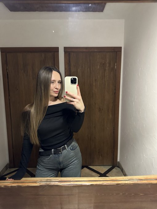 A woman takes a mirror selfie in a black off-shoulder top and gray jeans, standing in front of wooden doors.