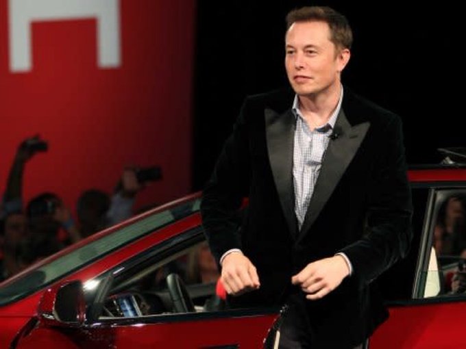 Elon Musk, wearing a black velvet tuxedo jacket and light blue shirt, steps out of a red Tesla vehicle at what appears to be a public event.