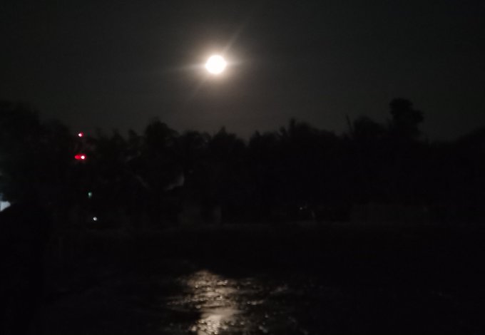 A nighttime photograph of a bright full moon hanging over a dark silhouette of trees, with light reflecting on water below.