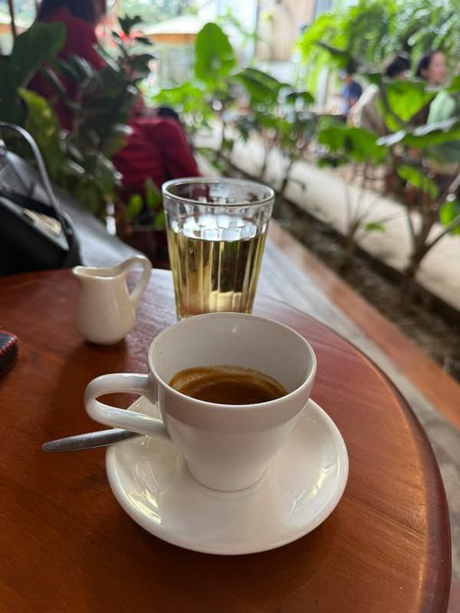 A cup of espresso and a glass of tea sit on a wooden table at an outdoor café.