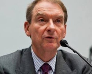 A close-up of an older man with grey hair in a suit and red tie speaking into a microphone, possibly in a formal hearing or press setting.