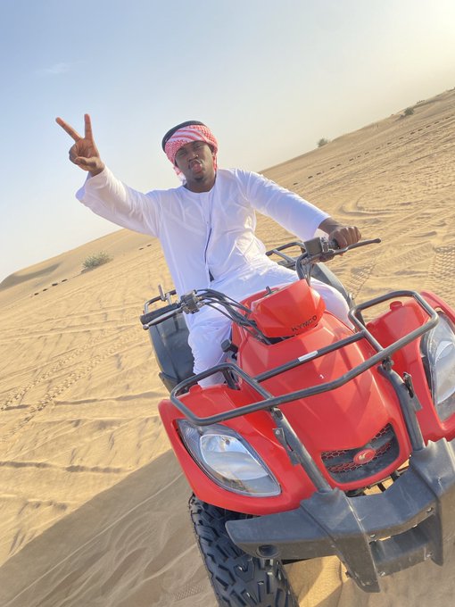A man in traditional white Arab attire and a red-and-white keffiyeh rides a red ATV through a desert, making a peace sign with his hand.