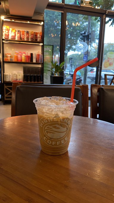 An iced coffee drink from "Highland Coffee" in a clear plastic cup with an orange straw, sitting on a wooden table in a café.