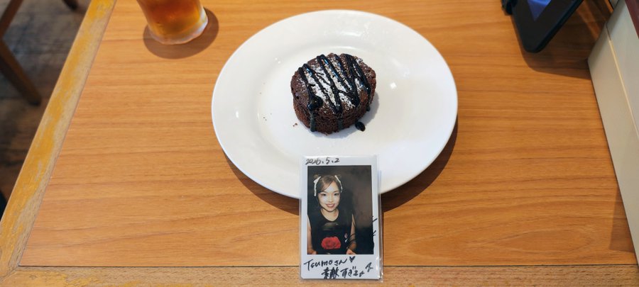 A small chocolate cake with chocolate drizzle sits on a white plate on a wooden table, with a Polaroid photo of a woman labeled 'Temmyson' and dated '2024.5.2' placed beside it.