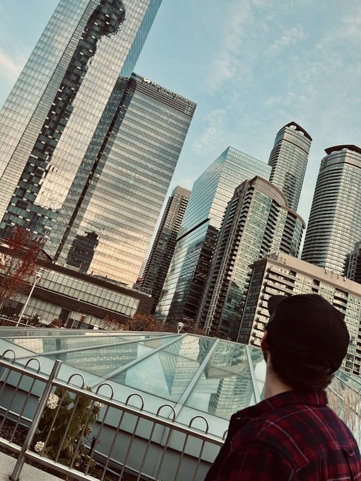 A person in a black beanie and red plaid shirt looks out over a glass railing at modern skyscrapers in a downtown city under a partly cloudy sky.