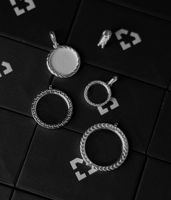Product photo of silver pendants and charms arranged on a black surface with white geometric logos, showing various ring-shaped designs including one with a twisted rope edge and another with a Greek key pattern.