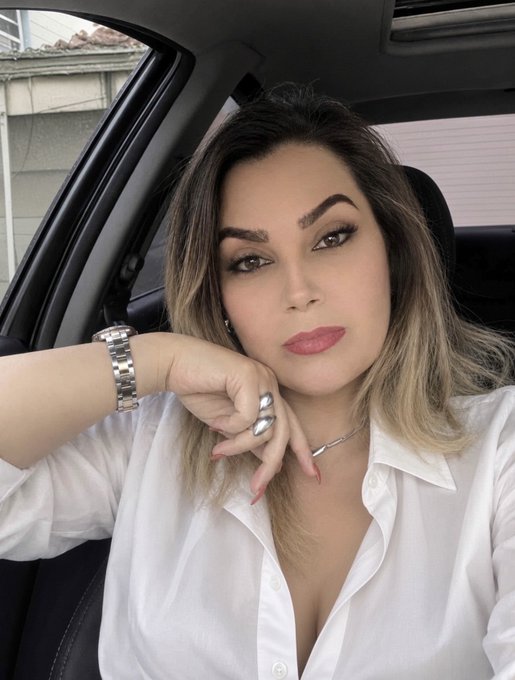 A selfie of a woman with dark hair wearing a white collared shirt, sitting in a car.