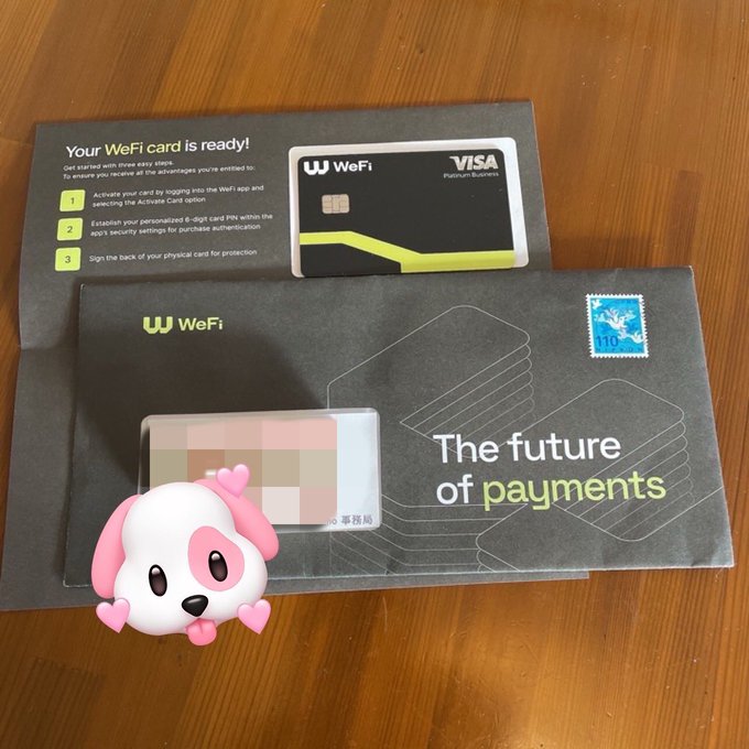 A WeFi Visa Platinum Business card sits on top of its "The future of payments" packaging on a wooden table, with a cute pink dog sticker covering part of the card details. The image shows the user receiving a new payment card, which loosely connects to the post about Japanese cryptocurrency exchanges tightening withdrawal restrictions and alternative ways to use crypto without converting to yen domestically.