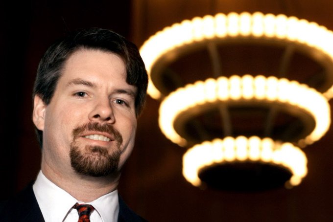 The image features a man wearing a suit and tie standing next to a large chandelier with multiple lights. The man appears to be smiling for the camera while posing near the lighted fixture, which adds an elegant touch to the scene. The background of the photo is dark, making it stand out even more. The caption provided seems unrelated to the main subject matter and does not provide any relevant information about the image or its context.
