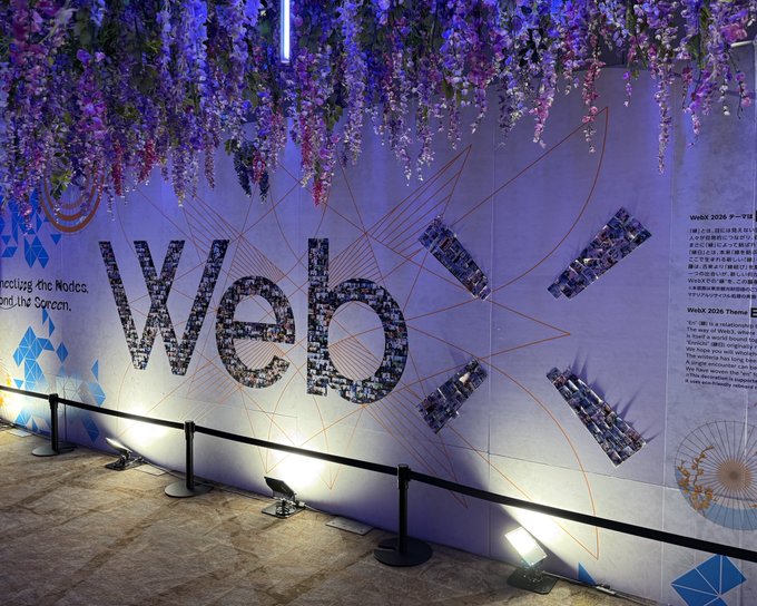 An event display wall spelling 'WebX' in large mosaic letters with red geometric lines, decorated with hanging purple flowers and spotlights, with explanatory text about WebX 2026.