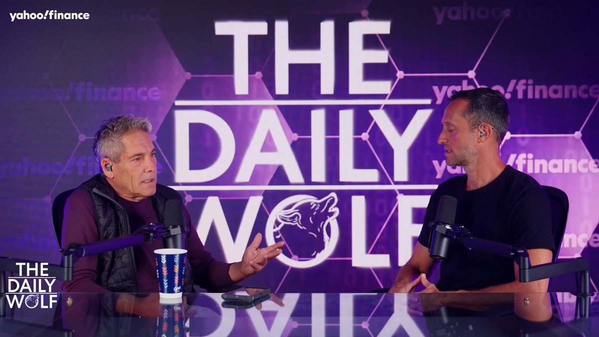 The image features a podcast interview between two men, with one man wearing a purple shirt and the other wearing a black shirt. They are sitting at a table, engaged in conversation about Bitcoin. The background of the image includes text that reads "Daily Wolf" and "Yahoo Finance," suggesting that this is an online platform for discussing financial topics. Additionally, there is a cup placed on the table between them.