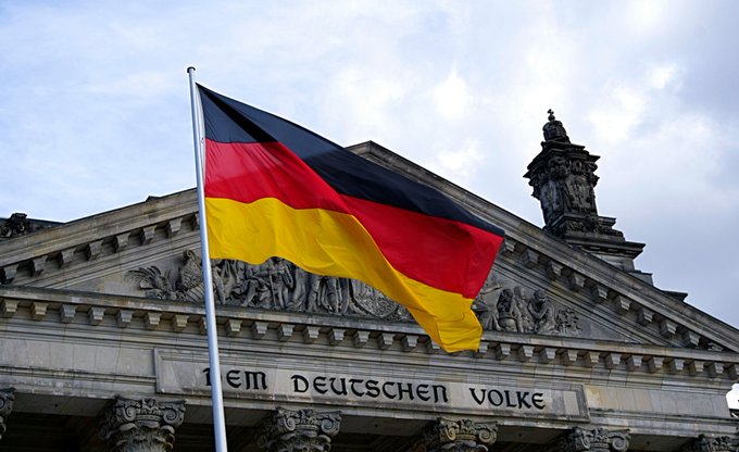 A German flag waves in the foreground in front of the Reichstag building, where the inscription "DEM DEUTSCHEN VOLKE" (To the German People) is visible on the facade, symbolizing the nation's recent legislative action offering 0% capital gains tax on Bitcoin held over one year.