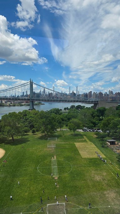 The image shows a soccer field with several people playing soccer, surrounded by trees and a bridge in the background. There are at least 12 visible players scattered across the field, engaged in various activities such as kicking the ball or running around. The scene is set against a backdrop of a city skyline, including a tall building that appears to be part of the New York City skyline. This image seems to be related to an event like theETH Conference held at the University of Edinburgh, where people gather for sports and social activities in the beautiful surroundings of the city.