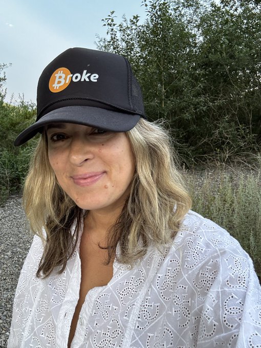 A woman wearing a white eyelet blouse poses outdoors in front of greenery, sporting a black trucker hat with an orange Bitcoin logo and the word "Broke."
