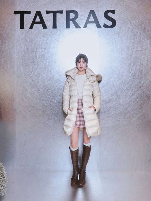 A young woman models a long, cream-colored puffer coat with a fur-trimmed hood, paired with a plaid skirt and tall brown boots, standing in front of a silver textured wall displaying the 'TATRAS' logo.