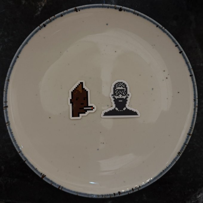 The image features a white plate with two stickers on it, one of which depicts a man smoking a cigarette while wearing a beard and glasses, and the other showing a man without any facial hair or glasses. The text "2017 x 2026 My CryptoPunk x my Normie" is also present in the image.