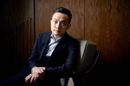 A man in a dark blue suit and light blue shirt sits in a chair against a wood-paneled wall, looking toward the camera.
