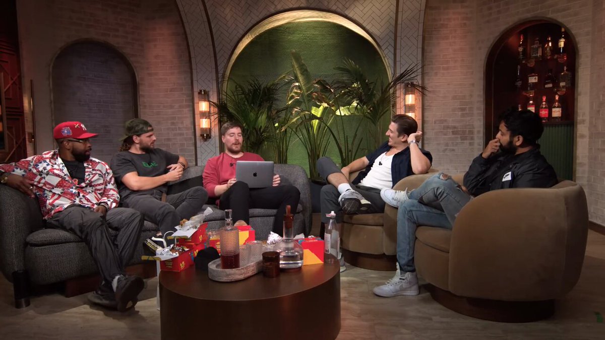 The image shows five men seated in a lounge-like podcast studio with arched brick walls, tropical plants, and a bar display in the background, gathered around a coffee table with McDonald's bags and drinks. This appears to be a still from MrBeast's podcast, where he and guests—including one holding an Apple laptop—discuss how he foolishly left his Bitcoin private key labeled on his laptop, resulting in a robbery and $2 million loss.