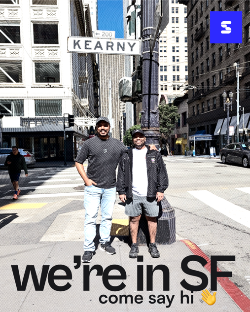Two men posing on a San Francisco street corner beneath a "KEARNY" sign, with a blue "S" logo and text reading "we're in SF come say hi" with a waving emoji.
