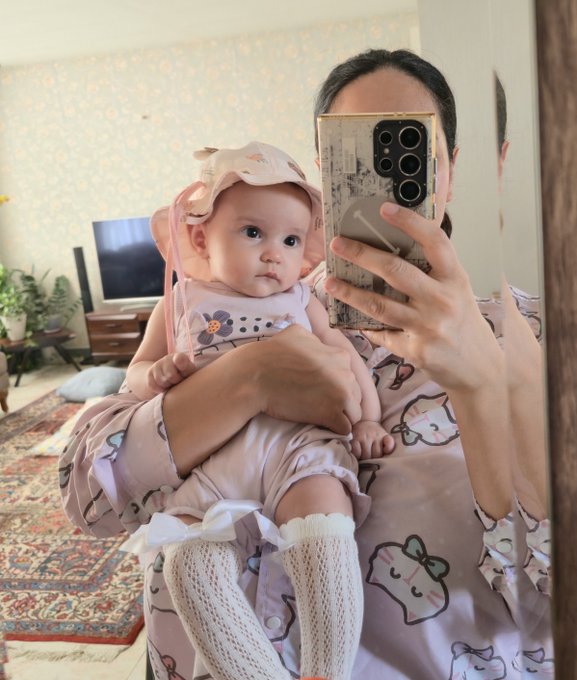A mirror selfie of a mother wearing pink pajamas holding a baby dressed in a pink outfit with a matching sun hat and white leg warmers, in a living room setting.