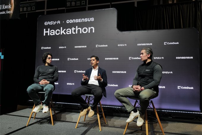 Three people seated on stools on a stage during an EASYX × Consensus panel discussion about a Hackathon, with a backdrop featuring CoinDesk, EASYX, and Consensus logos.