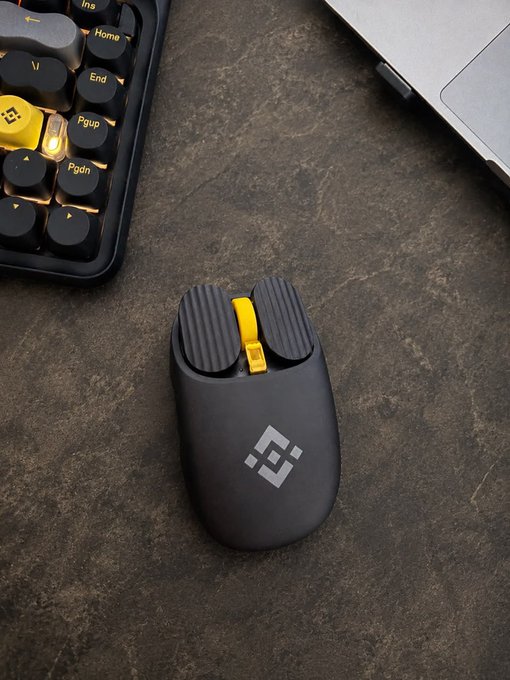 A black wireless mouse with a yellow scroll wheel and a white logo resting on a dark textured surface next to a mechanical keyboard and a laptop.