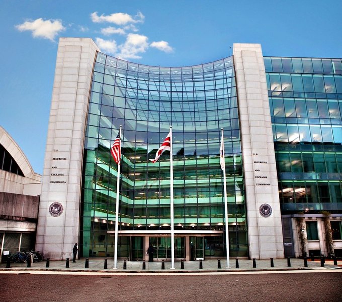 The U.S. Securities and Exchange Commission building featuring its modern curved glass facade, three flagpoles with the American flag and SEC flags, and official seals on the stone pillars.